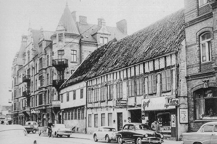 Äldre svartvitt foto i stadsmiljö med fina hus, bilar och fotgängare.