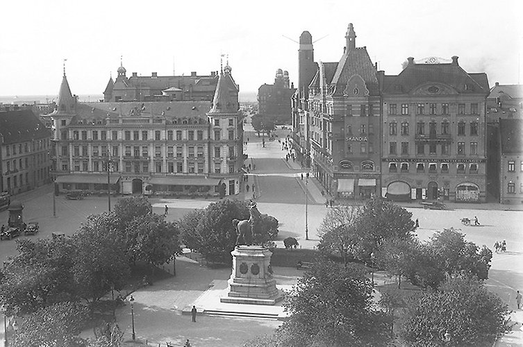 Äldre svartvitt foto i stadsmiljö med pampiga hus och ett torg med stor staty i mitten. 