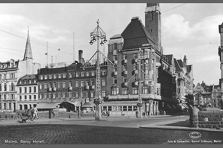 Äldre svartvitt foto i stadsmiljö med hus och ett torg i förgrunden. 