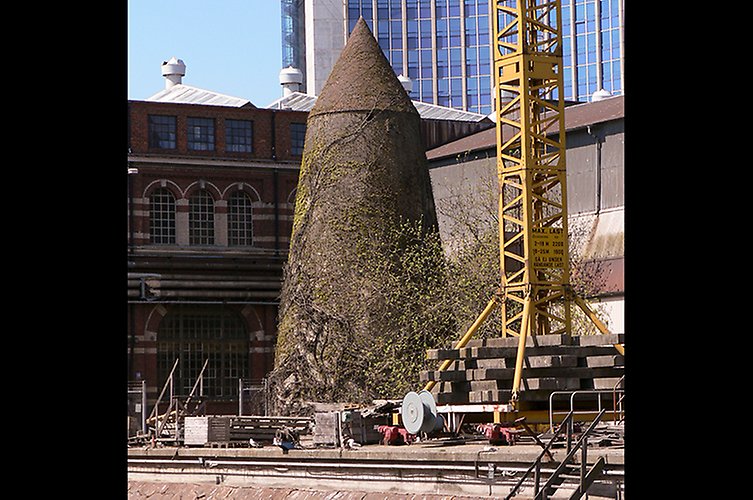 Märklig spetsig byggnad omgiven av äldre industribyggnader och en lyftkran. 
