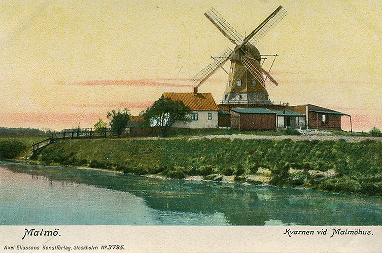 Äldre vykortsbild av väderkvarn med omgivande hus. Vatten i förgrunden. Solnedgång.