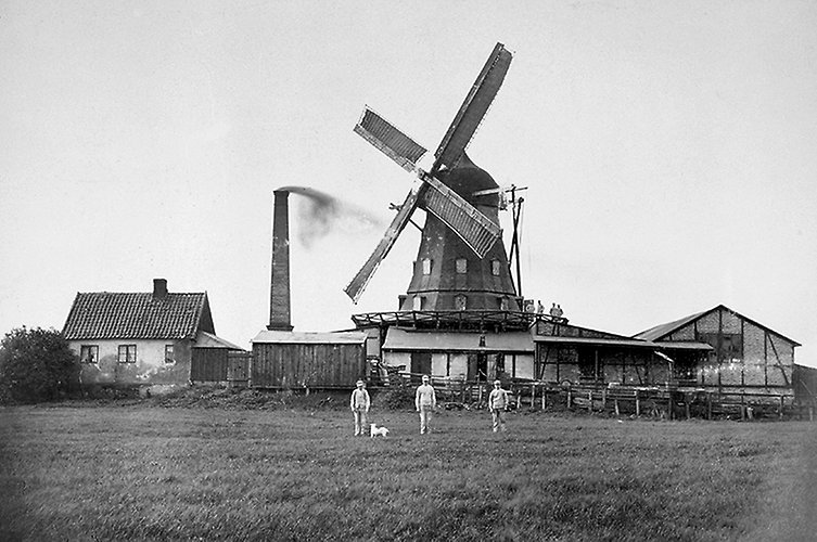 Äldre svartvitt foto på väderkvarn och omgivande hus. Ett fält i förgrunden med tre män som står framför väderkvarnen.