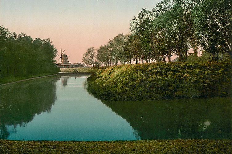 Äldre vykortbild med kanal i parkmiljö och väderkvarn i bakgrunden.