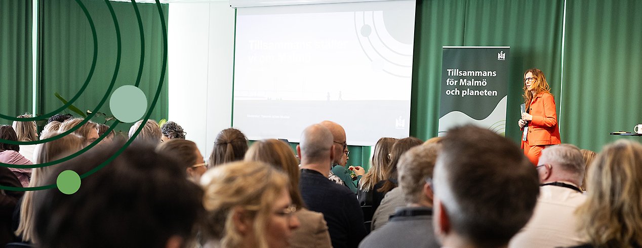 En kvinna i orange kostym står och pratar framför en fullsatt scen.