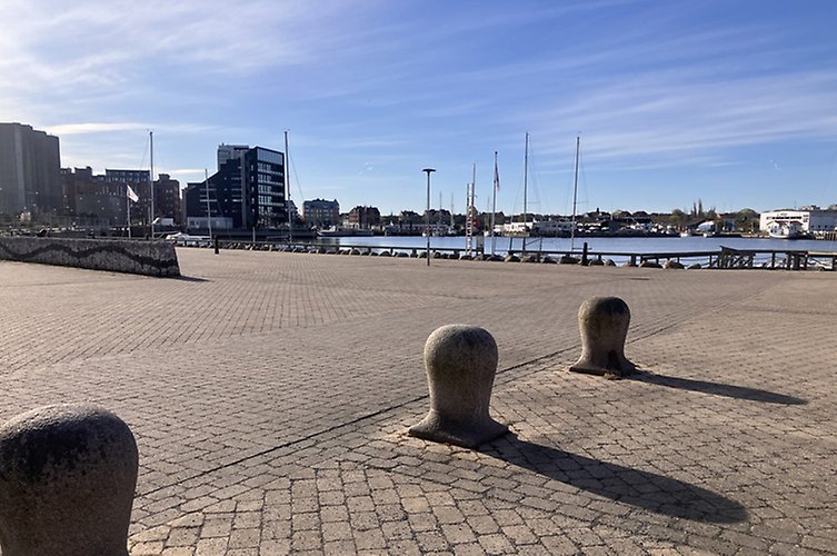 Offentlig plats med öppen stenlagd yta vid småbåtshamn. 