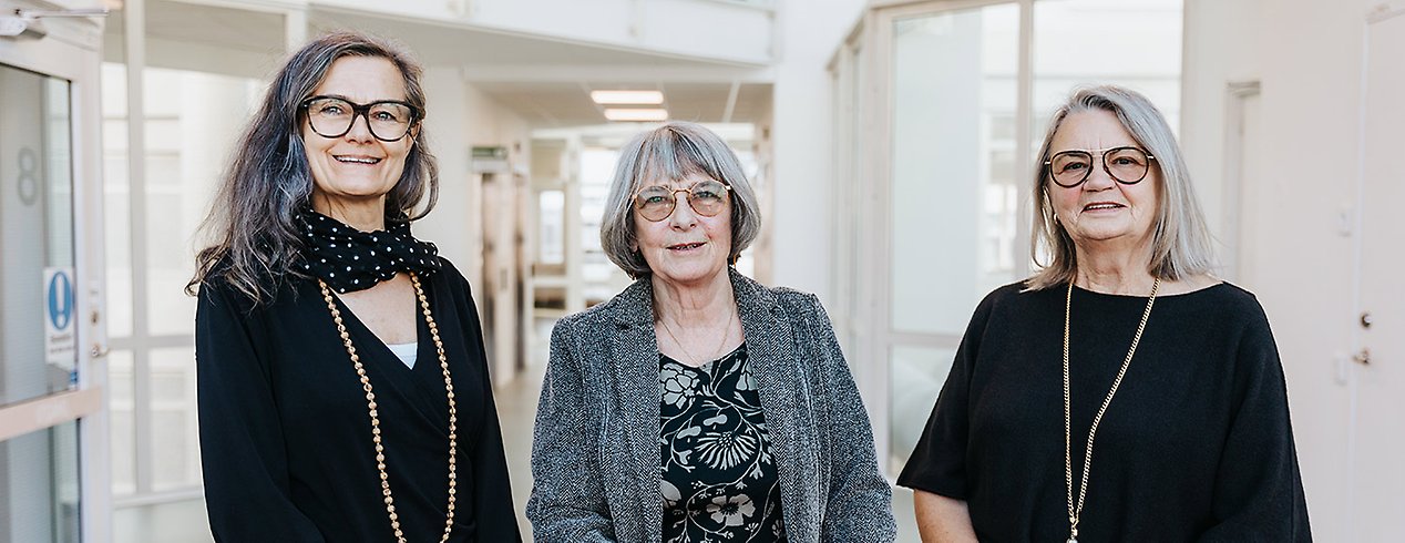 Marie-Louise, Pilka och Lisbeth står i en ljus vit korridor. De är alla mörkklädda, har glasögon och ett vänligt leende.