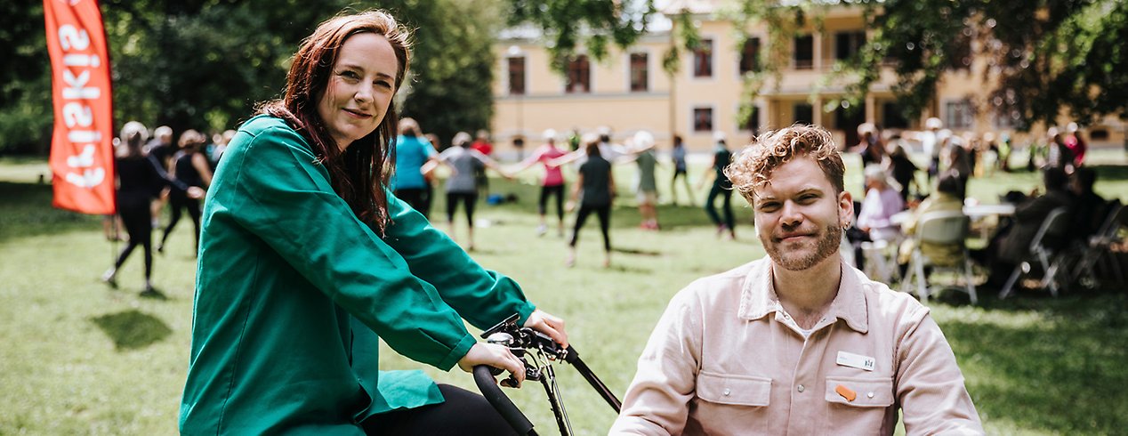 Freja och Adam sitter på och i en lådcykel. I bakgrunden syns jympande seniorer.