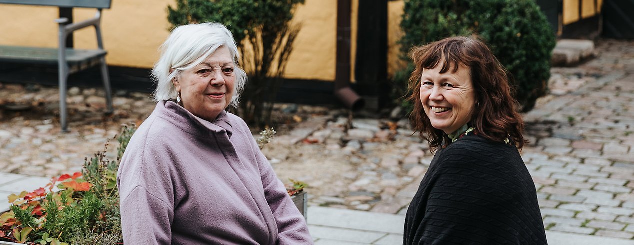 Ulla-Britt har grått hår och en lila tröja, Camilla har brunt hår och en svart tröja. De sitter på en bänk på en stenbelagd innergård.