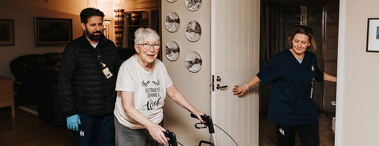 Mona öppnar dörren för Ingbritt och hennes rollator medan Hashem följer efter bakom.