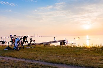 Två cyklar står nära en brygga vid Sibbarpstranden där flera personer går ner i vattnet. I bakgrunden kan man se solnedgången och Öresundsbron. 