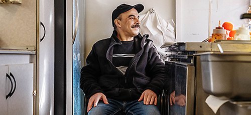 En man med mustasch sitter på en stol inne i en falafelvagn. Han ser ut mot fönstret och ler. Han har keps och svart jacka.