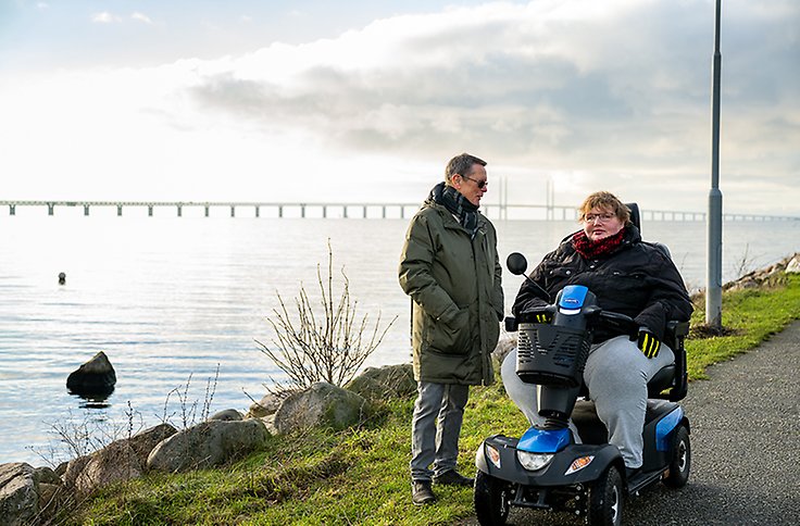 En kvinna i rullstol bredvid en man som står upp och tittar på henne. De har på sig tjocka jackor, i bakgrunden syns havet och Öresundsbron.