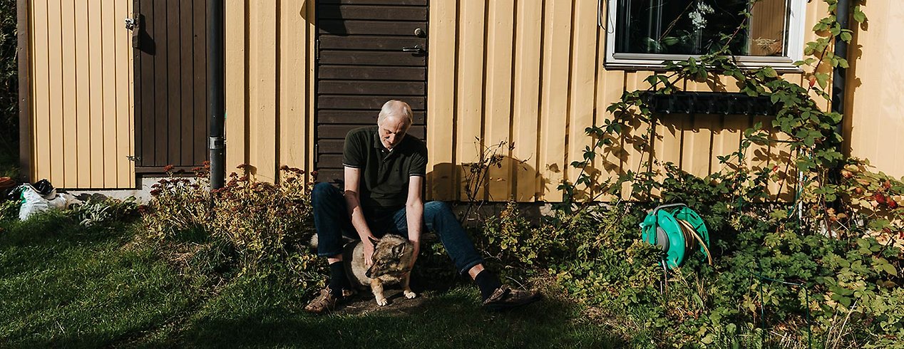 Sven sitter framför sitt hus med hunden framför sig. Huset är omringat av vilda buskar.