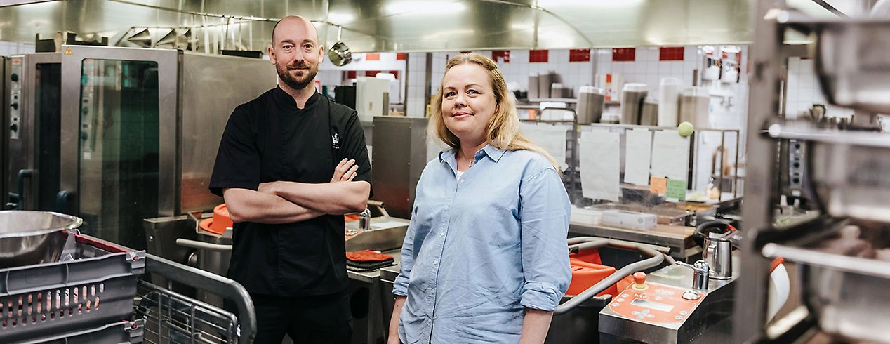 Joakim och Agneta står i Mathildenborgs kök. Joakim har svart klädsel med armarna i kors. Agneta har blont hår och en blå skjorta.