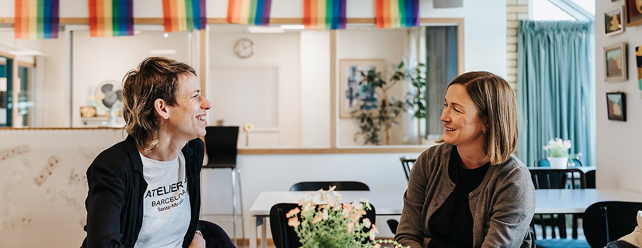 Emilie och Christina sitter på en mötesplats, i bakgrunden hänger en regnbågsflagga.