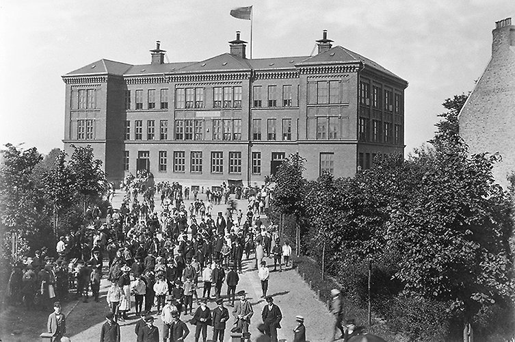 Gammalt svartvitt foto på skolbyggnad. Mycket folk utanför skolan. Flaggning.