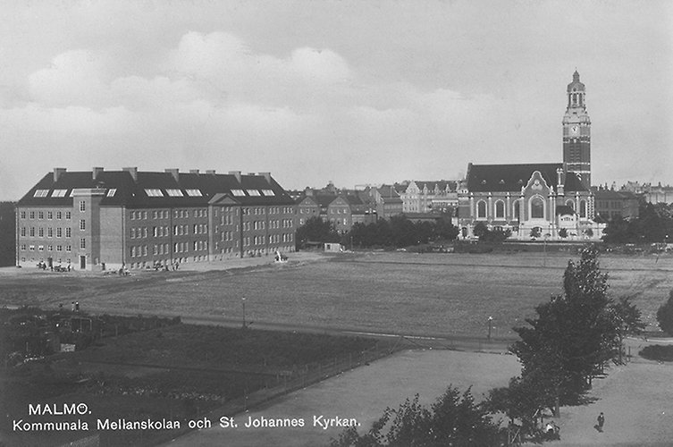Gammalt svartvitt foto med vy över större tegelbyggnad, kyrka och fält.