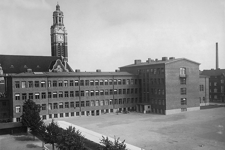 Äldre svartvitt foto på skolbyggnad i tegel. Kyrktorn i bakgrunden.