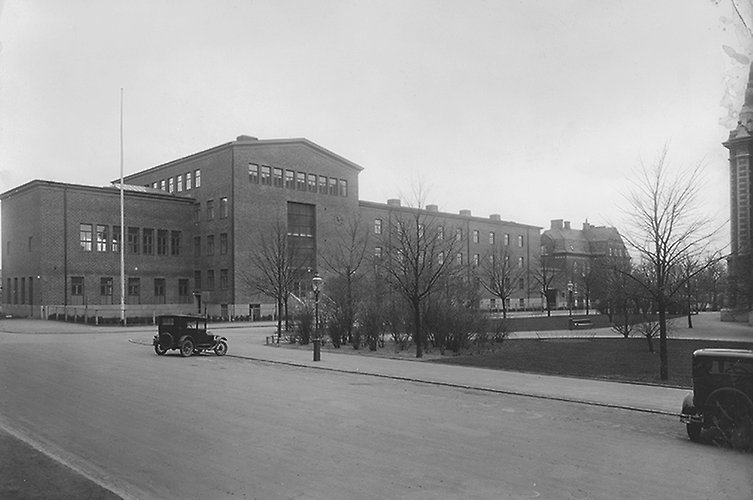 Äldre svartvitt foto på skolbyggnad i tegel. Tidsenliga bilar i förgrunden.