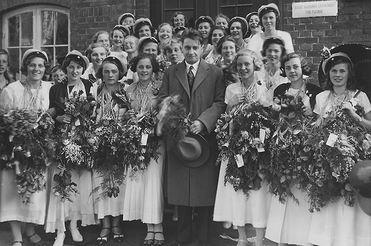 Runt 20 flickstudenter med blommor poserar utanför byggnad. Svartvitt foto.