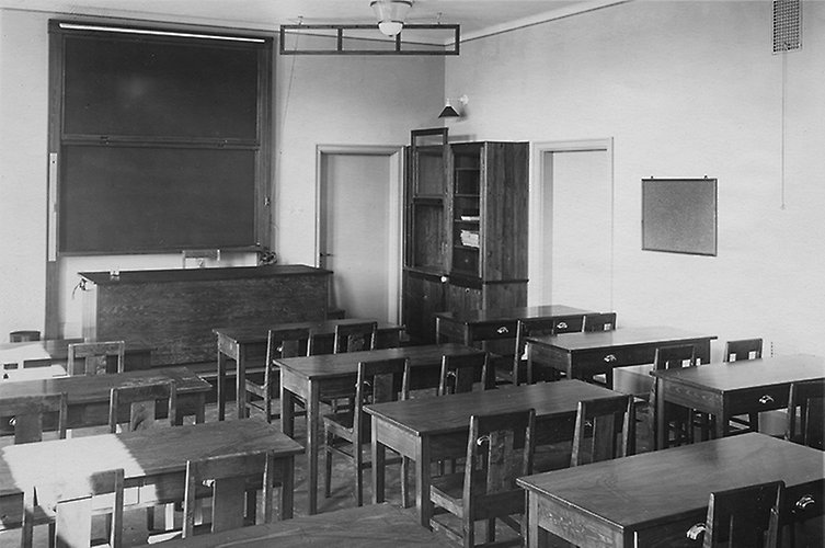 Äldre svartvitt foto från klassrum med skolbänkar, kateder och griffeltavla.