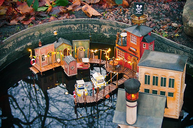 Miniatyrhus i hamnmiljö med hus, brygga och en liten fyr, uppbygd och flyter i en fontän. 