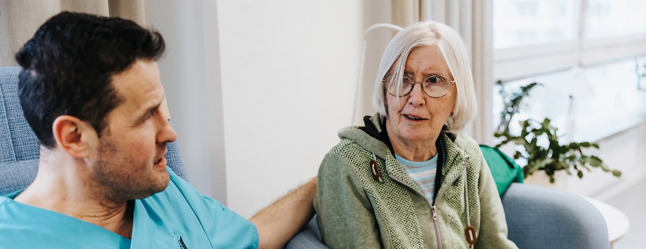 En man och en äldre kvinna sitter i en blå soffa. Han har handen på hennes rygg.