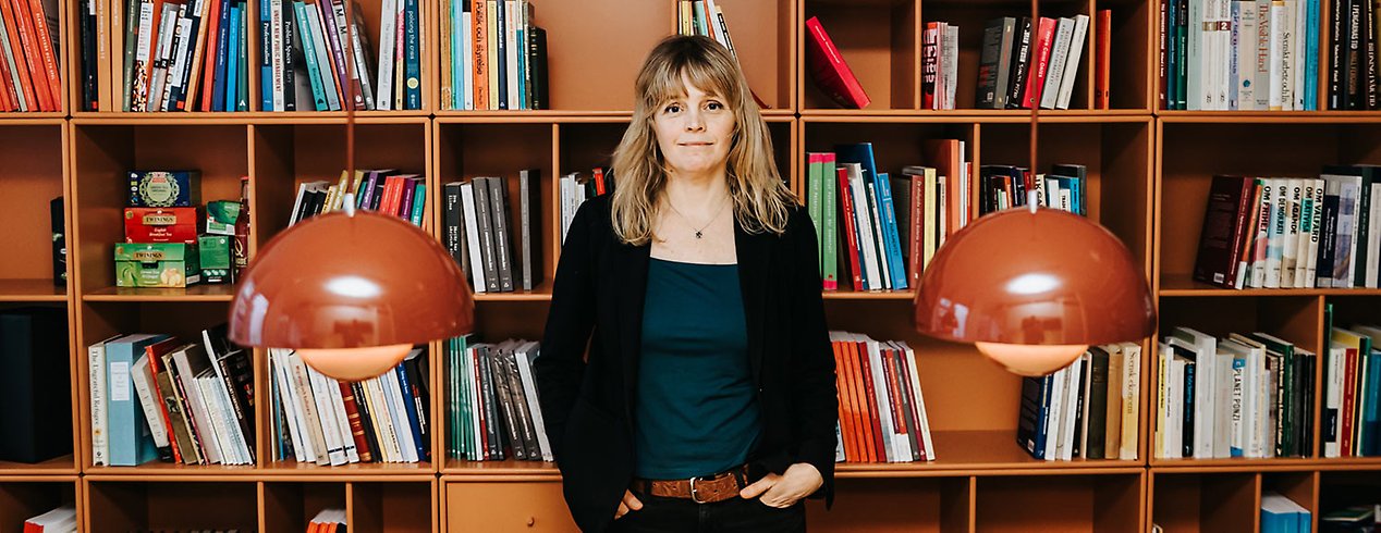 Kvinna med blont långt hår står lutad mot en bokhylla.