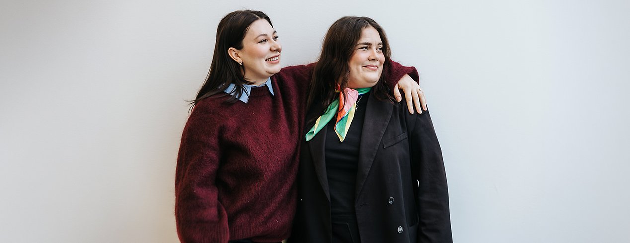 Två kvinnor, den ena med armen om den andras axlar. Båda ler och ser åt sitt vänster.