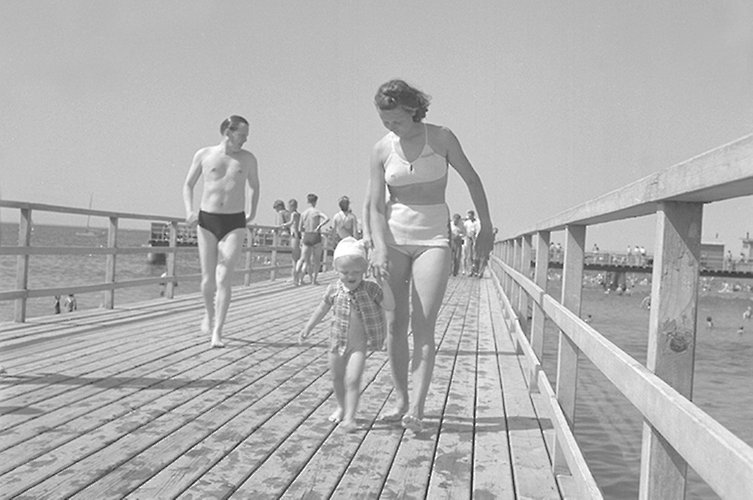 Äldre svartvitt foto. Pir med badhus i bakgrunden. Badklädda fotgängare. 