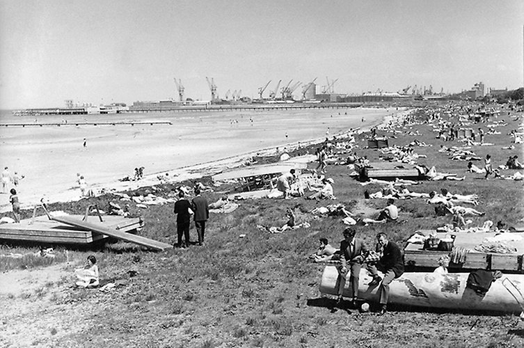 Strand och många badgäster. Fabriker i bakgrunden. 
