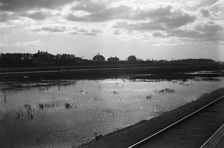 Äldre svartvitt foto. Järnväg i förgrunden, bebyggelse på andra sidan en vik. 