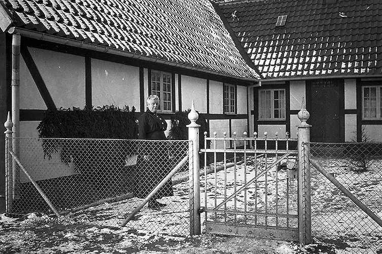 Gammalt svartvitt foto. En kvinna står utanför ett korsvirkeshus. Snö på marken.