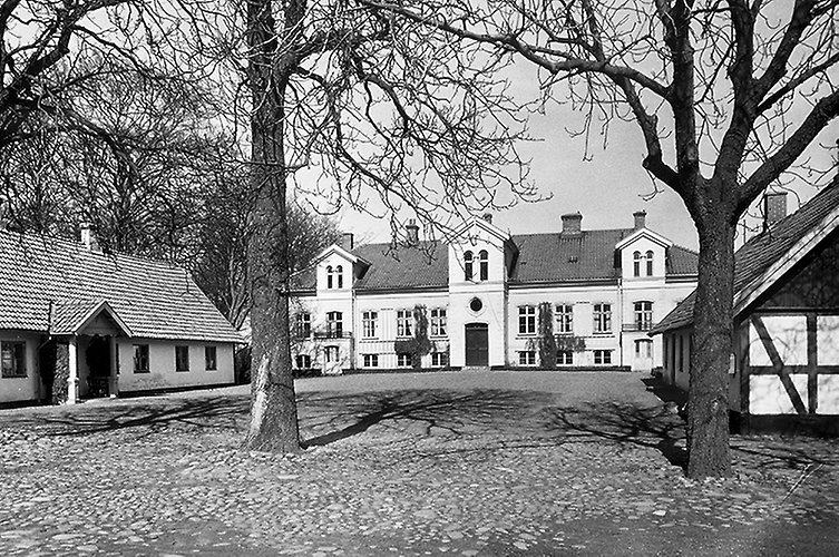 Gammalt foto. Herrgård och tillhörande hus med en tom gårdsplan i förgrunden.