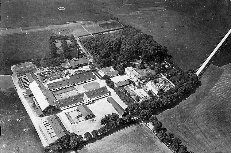 Gammal svartvitt flygfoto över ett 20-tal byggnader på en åker.