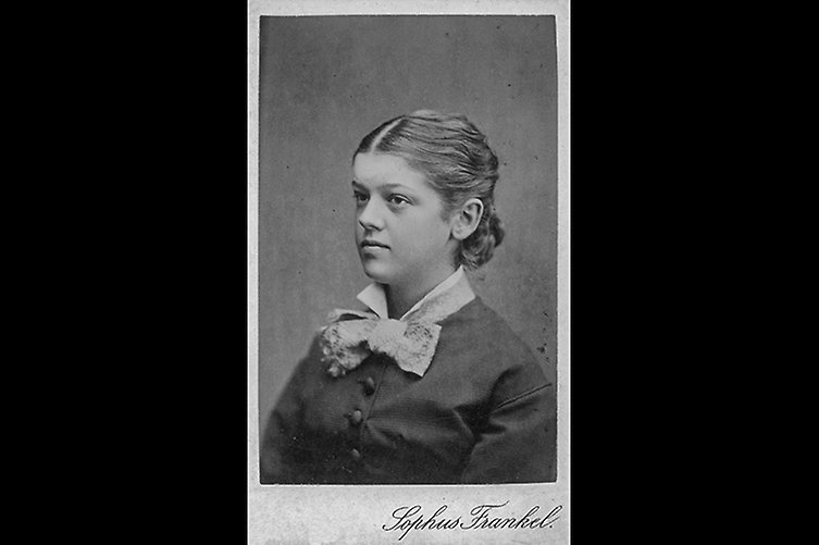 Gammal svartvitt porträtt på ung kvinna med uppsatt hår och fina kläder.