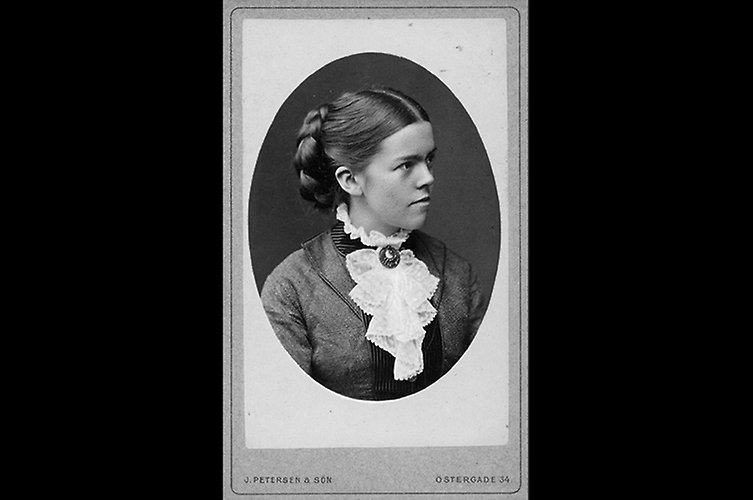 Äldre svartvitt porträtt av ung kvinna med uppsatt hår och fina kläder.