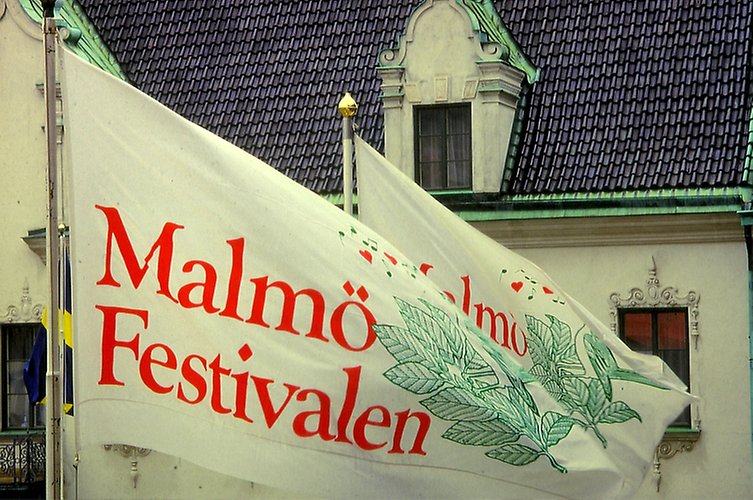 Bild på flaggor med festivalens tidigare logga. Flaggorna är vita med "Malmö Festivalen" skrivna i röd text. På bilden syns även kvistar målade i grönt. 
