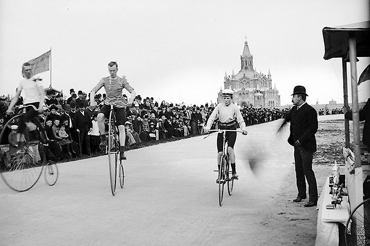Tre cyklister på bana inför publik och kyrka i bakgrunden. Svartvitt foto.