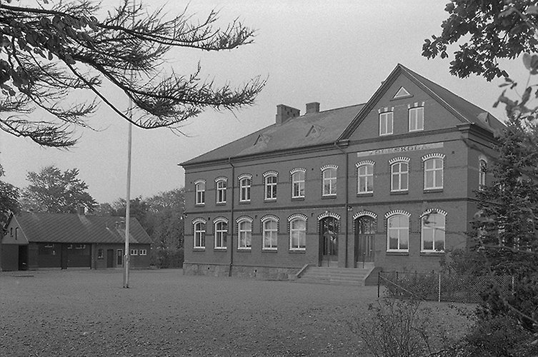 Äldre skolbyggnad i två våningar, öde skolgård en flaggstång. 