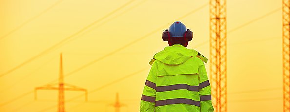 Elektriker står i ett fält med elmaster och tittar bort mot solnedgången.