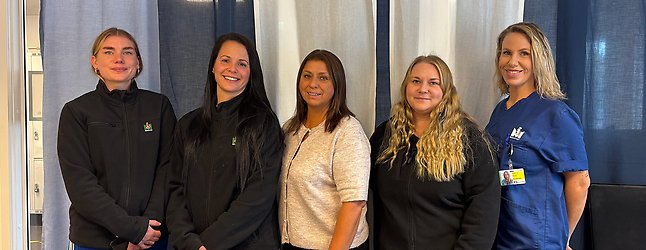 Fem kvinnliga undersköterskor står på en rad och tittar mot kameran. Några har sina arbetskläder och några har sina privata kläder. 